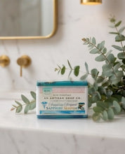 A rectangular bar of bath soap from Bath Essentials, An Artisan Soap Co., labeled 'Sapphire Serpent', placed on a white marble surface with eucalyptus leaves and a gold-framed mirror in the background.