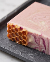 A close-up of a rectangular handmade soap bar with a pink and cream marbled swirl pattern and a honeycomb cell design on one end, resting on a dark slate tray.