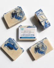 Four artisan soap bars with blue and cream swirls are arranged around a wrapped soap package on a clean white background. The soap has a warm cream base with bold blue and dark navy marbled patterns.