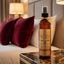 A bottle of RDCG Essentials Green Collection 'Stay While' body and skin spray, placed on a marble bedside table in a warmly lit bedroom setting with a bed and pillows in the background.