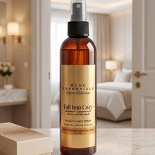An amber glass room spray bottle with a black spray top and a gold label reading 'RCDC Essentials Home Collection Fall Into Cozy' sits on a light marble surface in a softly lit bedroom setting. A matching cream gift box is partially visible at left.