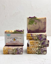 Four artisan soap bars with a white, purple, green, and gold marbled pattern, including two stacked bars and two standing bars, with one bar in front showing its paper label that reads 'Lavender & Honey' by 'Puck Essentials'.