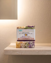 Two artisan soap bars stacked on a light stone shelf against a warm beige wall. The top bar has a white paper label reading 'Red Earth Essentials An Artisan Soap Co.