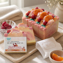 A photo of bars of handmade soap decorated with raspberry and tangerine designs, with a slice of the bar showing the swirled pink and white colors. The soap is accompanied by decorative flowers and the packaging is visible.