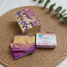 Three soap bars with purple and yellow swirls on a woven mat with a box of Artisan Soap Co.