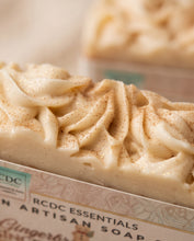 A close-up of a piped bar of artisan soap with a cream-colored body and fine brown spice specks, resting on a branded RCDC Essentials paper sleeve. The background is softly blurred with another soap bar visible.