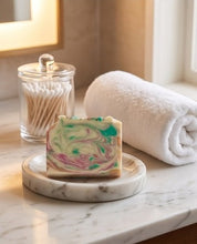 A rectangular bar of marbled soap with swirls of green, pink, and cream sits on a round marble dish on a bathroom counter, with a rolled white towel and a glass jar of cotton swabs in the background.