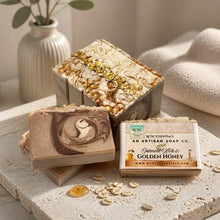 Two artisan soap bars and their packaging boxes arranged on a stone surface with oats and a vase in the background. The front box reads 'Rose Essentials An Artisan Soap Co.