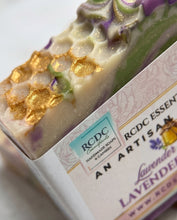 A close-up of a handmade lavender soap bar with gold foil accents and embedded botanical details, partially wrapped in a paper sleeve labeled 'RCDC ESSENTIALS' and 'Lavender'.