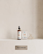 Two DW Bath & Body Works products, a dark amber spray bottle and a clear pouch with a white label, displayed on a white shelf against a textured beige wall, with a small sign below reading 'New Collection'.