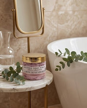A jar of whipped soap scrub sits on a marble side table in a bathroom setting, next to a white bathtub, a gold-framed mirror, and green eucalyptus leaves.