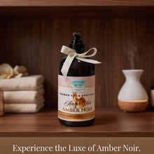 A bottle of RCD Cessation Shower Gel & Body Wash in Amber Noir scent, styled on a wooden shelf with a diffuser and towels in the background. The bottle has a black pump and collar, a cream ribbon bow, and a label with amber, pink, and white tones.