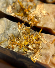 Close-up of a textured stone tray or dish with dried yellow-orange botanicals and scattered gold leaf flakes on a warm beige surface.