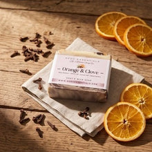 A rectangular bar of soap labeled 'Orange & Clove' sits on a folded cream cloth on a rustic wooden table, surrounded by dried orange slices and cloves. The brand name 'Good Essentials' is visible on the label.