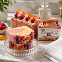 A photo of bars of handmade soap decorated with raspberry and tangerine designs, with a slice of the bar showing the swirled pink and white colors. The soap is accompanied by decorative flowers and the packaging is visible.