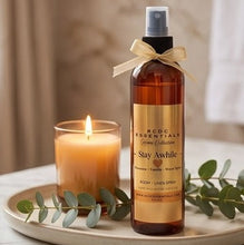RCDC Essentials Home Collection Stay Awhile room and linen spray in an amber bottle with a gold label and ribbon, styled on a tray next to a lit candle with eucalyptus leaves.
