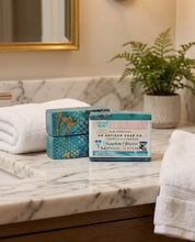 Two rectangular bars of soap from An Artisan Soap Co. displayed on a white marble bathroom counter next to folded white towels, with a potted fern in the background.