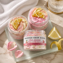 Jars of whipped soap sugar scrub with pink and yellow colors, alongside slices of lemon and pink flowers, indicating a citrus scent profile.