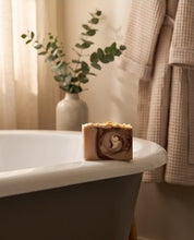 A rectangular bar of artisan soap with a brown swirl pattern sits on the edge of a white bathtub in a warm, softly lit bathroom. A beige robe hangs on the right, and a vase with green eucalyptus stems stands in the background.