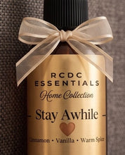 RCDC Essentials Stay Awhile home fragrance bottle with a black cap, a gold label, and a sheer beige ribbon bow, set against a dark textured background.