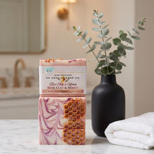 A bar of artisan soap from An Artful Soap Co. sits on a white marble countertop next to a black vase with eucalyptus and a folded white towel.
