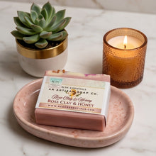 A bar of soap from Arctic Essentials sits on a pink marble dish on a white marble surface, styled with a small succulent in a gold pot and a lit amber candle.