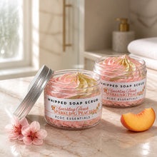 Jars of Sparkling Peach Whipped Soap Sugar Scrub, with a pink and white color scheme, displayed on a white surface with a bouquet of pink flowers in the background.