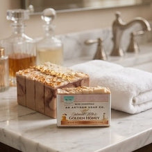 A bar of Johnnie Walker & The Artisan Soap Co. Golden Honey soap sits on a white marble bathroom counter next to a folded white towel, with glass bottles and a faucet blurred in the background.