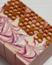 A close-up of a rectangular bar of handmade soap with a swirled pink and white pattern, embedded with purple glitter, topped with a textured golden-brown honeycomb design.
