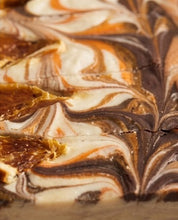 A close-up of a marbled dessert, likely a brownie or fudge bar, featuring swirls of dark chocolate, light brown, and creamy off-white with golden caramel-like accents.