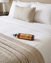 A bottle of hair product lies on a neatly made bed with white linens, a beige textured pillow, and a cream knit throw blanket.