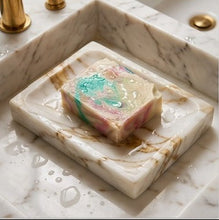 A rectangular marble soap dish with gold veining sits in a matching marble sink. A marbled soap bar with pink, teal, and cream swirls rests on the dish.