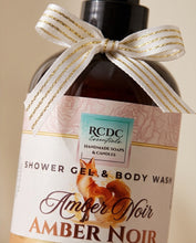 An amber glass bottle of RCDC Essentials Shower Gel & Body Wash in the Amber Noir scent, decorated with a white and gold striped ribbon bow, set against a warm beige background.