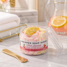 Jars of whipped soap sugar scrub with pink and yellow colors, alongside slices of lemon and pink flowers, indicating a citrus scent profile.