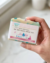 A hand holding a bar of handmade soap with a decorative paper label. The soap has a marbled cream, mint green, and blue color, and the label reads 'RCDC Essentials An Artisan Soap Co.