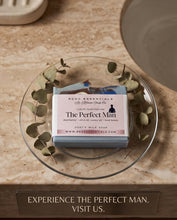 A bar of soap labeled 'The Perfect Man' by RCDC Essentials, wrapped in a white paper band and placed on a clear glass plate with eucalyptus leaves, set on a beige stone surface.