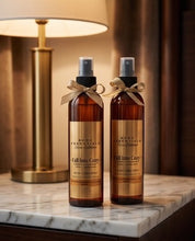 Two amber spray bottles of MCDS Essentials 'Fall Into Cory' hair care product with gold labels and beige ribbon bows, displayed on a marble countertop next to a lit table lamp.