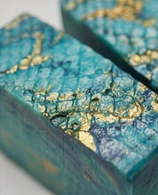 Close-up of a rectangular bar of soap with a textured turquoise and blue surface decorated with shimmering gold leaf flakes, set against a white background.