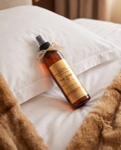 A bottle of body oil with a black spray top and a beige ribbon bow, resting on white pillows and a brown textured blanket.