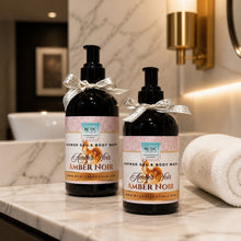 Two amber glass pump bottles of REDTC Amber Noir shower gel and body wash, tied with silver patterned ribbons, displayed on a marble bathroom counter next to a white towel.