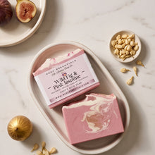 A styled flat-lay of a bar of soap in a pink and white marbled swirl, with its paper label reading 'Wild Fig & Pink Ashmore' by Soul Essentials. The soap sits in a beige ceramic dish on a white marble surface, surrounded by figs, pumpkin seeds, and dried flower petals.