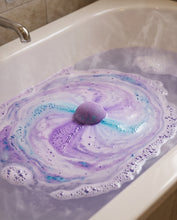 A purple bath bomb floating in swirling purple, blue, and white foam inside a white bathtub.