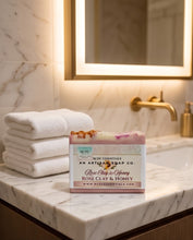 A rectangular bar of handmade soap labeled 'Rose Clay & Honey' by Redc Essentials, placed on a marble bathroom counter next to folded white towels and a gold faucet.