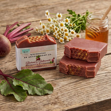 Two rectangular bars of beetroot and butternut soap with honeycomb tops are displayed on a rustic wooden table, alongside a wrapped bar, fresh beet greens and root, chamomile flowers, a jar of honey, and a wooden honey dipper. A dark red banner at the bottom reads 'SHOP THE C.