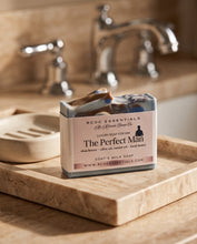 A bar of handmade soap labeled 'The Perfect Man' by RCDC Essentials sits on a marble soap dish on a bathroom counter with blurred faucets in the background.