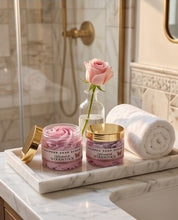 Two pink whipped soap bars in glass jars with gold lids sit on a marble tray on a bathroom counter, alongside a rolled white towel, a small vase with a pink rose, and a gold shower door in the background.