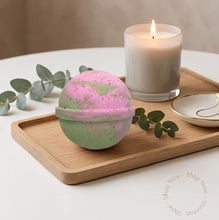 A round bath bomb with a pink and mint green marbled pattern sits on a wooden tray next to a lit candle, a small dish, and eucalyptus leaves.