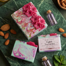 Two decorative handmade soap bars and their boxes are arranged on a green marble surface with almonds, a bottle, and mint leaves. The soaps have pink, mint green, and white marbled swirls, with one topped by large pink flower decorations.