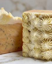 Close-up of handmade soap bars on a light marble surface, with one bar sliced to show a pale yellow and peach layer with dark speckles, and another bar featuring embossed floral star patterns in a creamy beige tone.