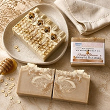 Three artisan honey and oat milk soap bars are arranged on a beige stone surface with a matching dish, folded cloth, honey dipper, oats, and the soap packaging box. The soaps have decorative swirls and embedded honeycomb details, with one bar topped by three bees.